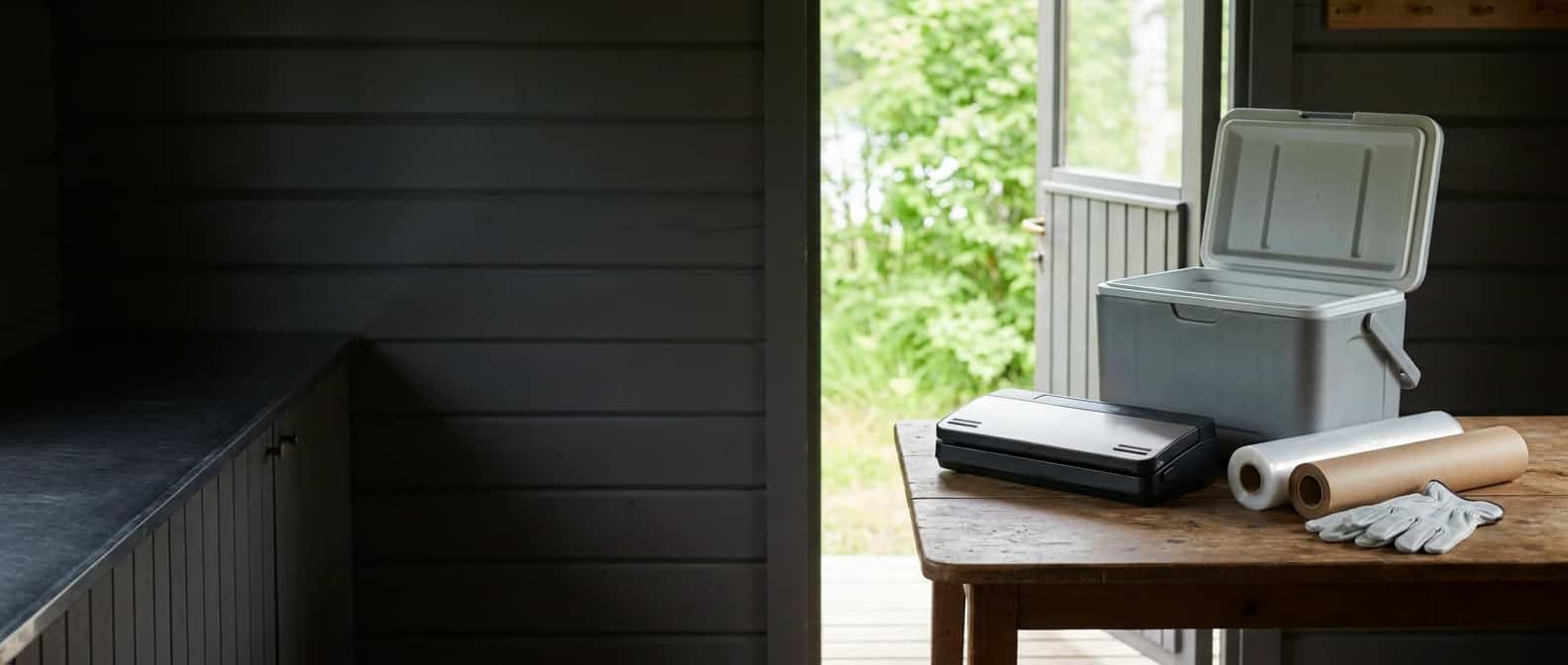 Vacuum sealer, cooler, freezer paper, and clean gloves are organized at a summer cabin workstation